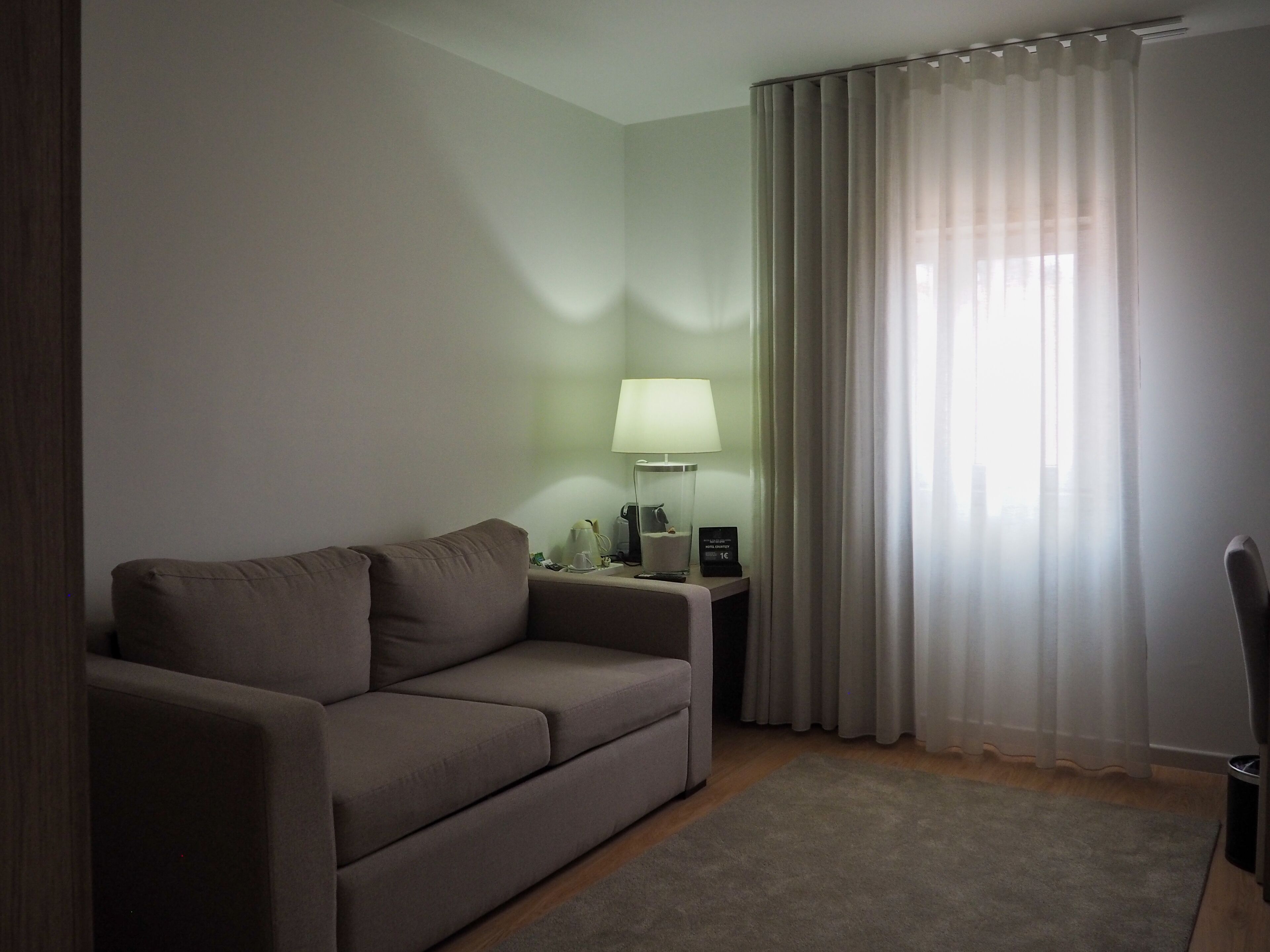 A modern hotel room with a grey sofa, a bedside table with a lamp, and sheer curtains.