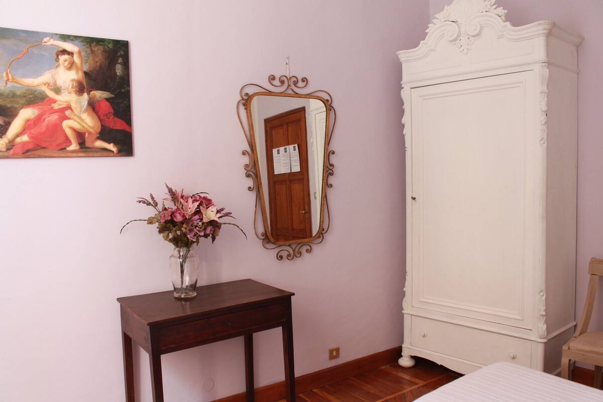 Een slaapkamer met een houten dressoir, een spiegel, een klein tafeltje met een bloevas en een bed.