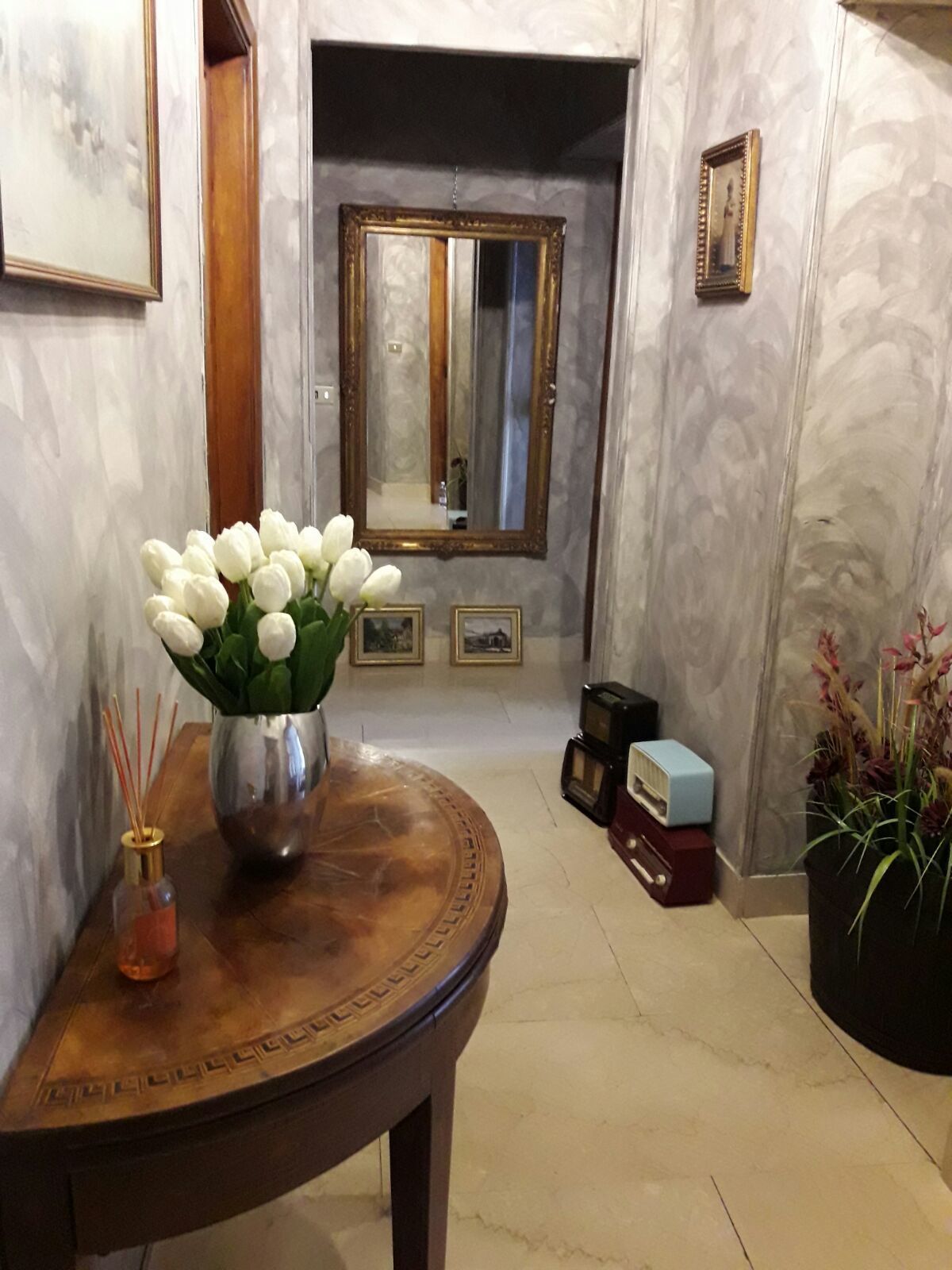 Een gang met een marmeren wand, een ronde houten tafel met een vaas witte bloemen, een spiegel en enkele ingelijste foto's.