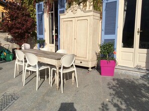Un espace repas extérieur avec une table en bois et des chaises blanches, un grand meuble et un pot de fleurs rose.