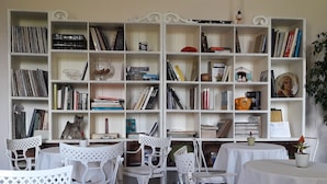 Een kamer met een grote witte boekenkast vol boeken en decoratieve objecten, witte stoelen rond een tafel, en een kleine potplant op de tafel.