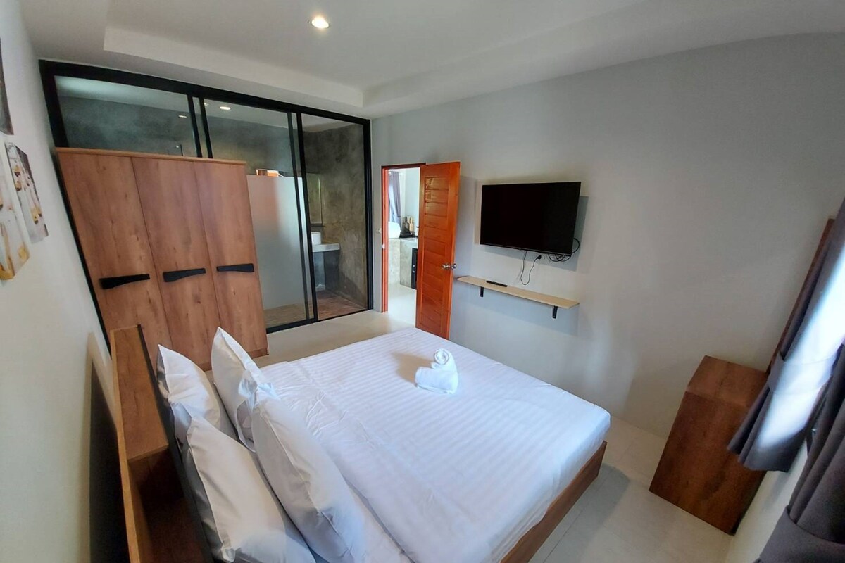 A modern bedroom with a large bed, wooden wardrobe, a TV mounted on the wall, and a sliding glass door leading to a bathroom.