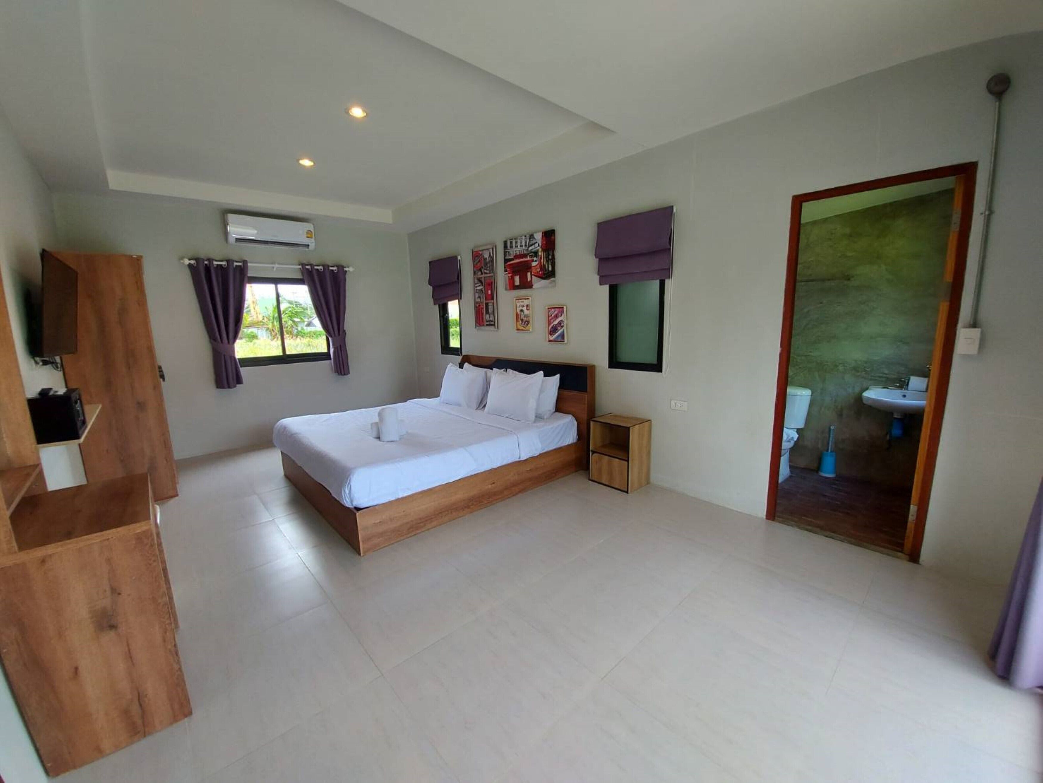 A bedroom with a bed, a bedside table, a window with curtains, a wall-mounted air conditioner, and a bathroom visible through an open door.