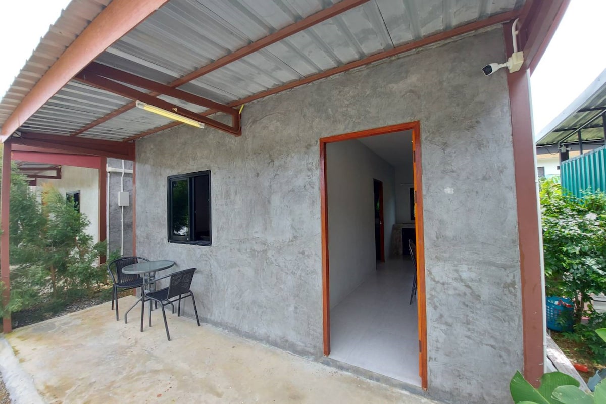 Uma área externa coberta com piso de concreto, uma mesa com cadeiras e uma porta de entrada para um espaço interno.