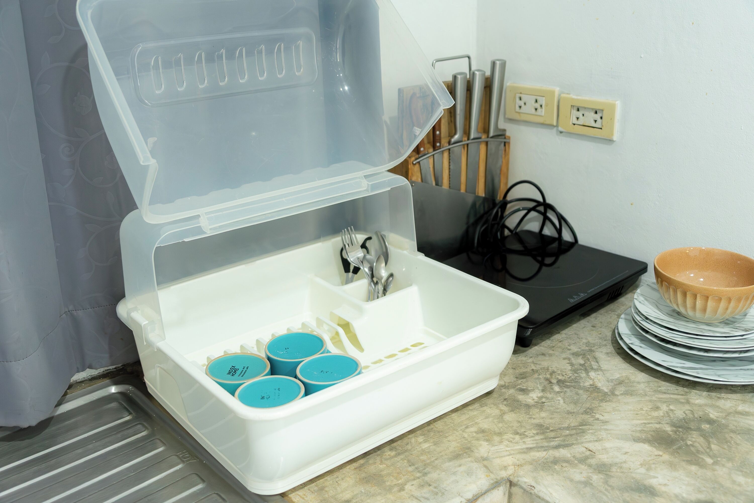 Un évier de cuisine surmonté d’un couvercle transparent, un ordinateur portable noir et une pile d’assiettes et de bols sur le plan de travail.