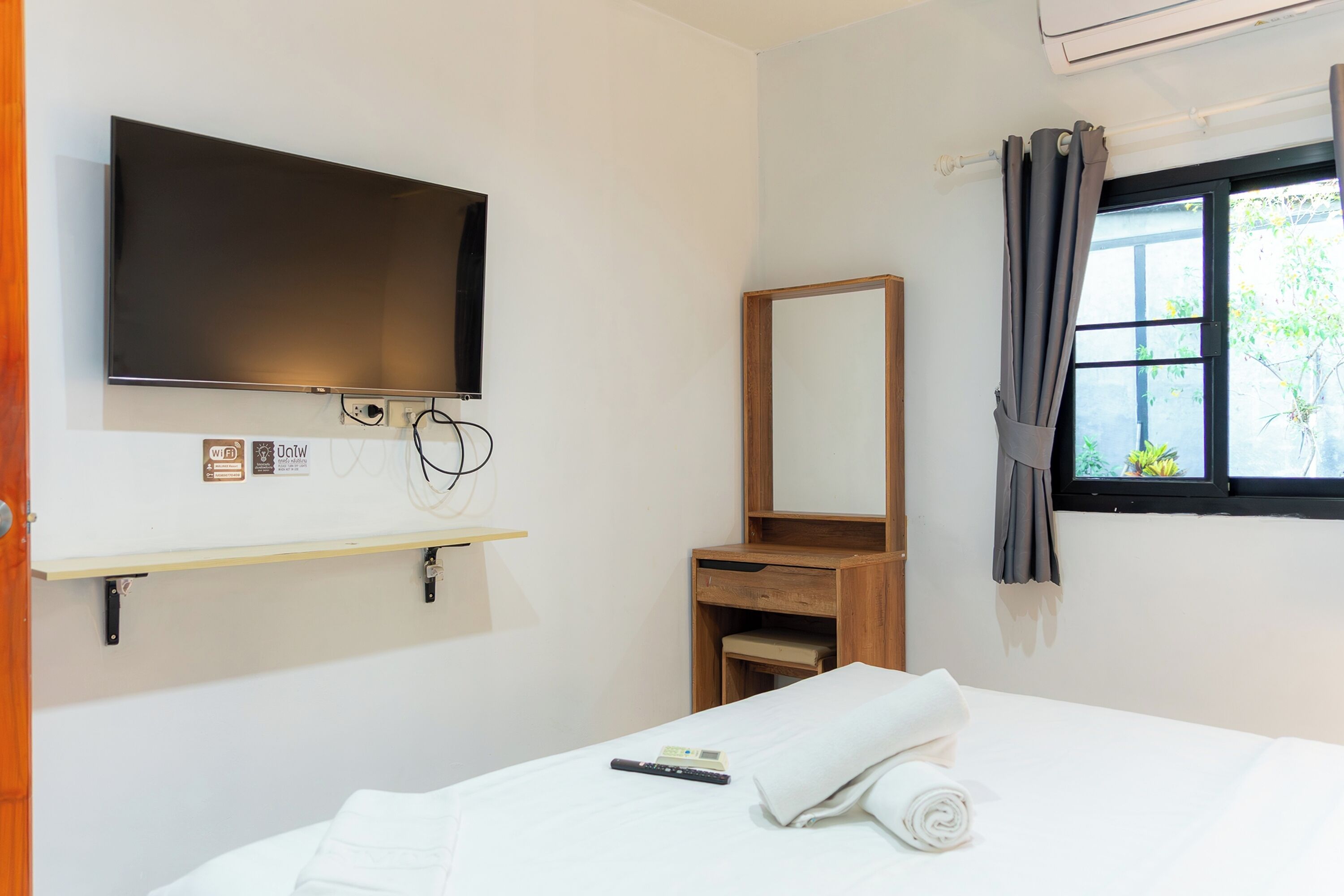 A hotel room with a bed, a TV mounted on the wall, a wooden dresser, a mirror, and a window with curtains.