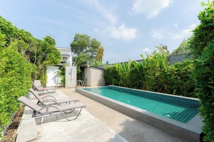 A swimming pool with lounge chairs, surrounded by greenery and a building in the background.