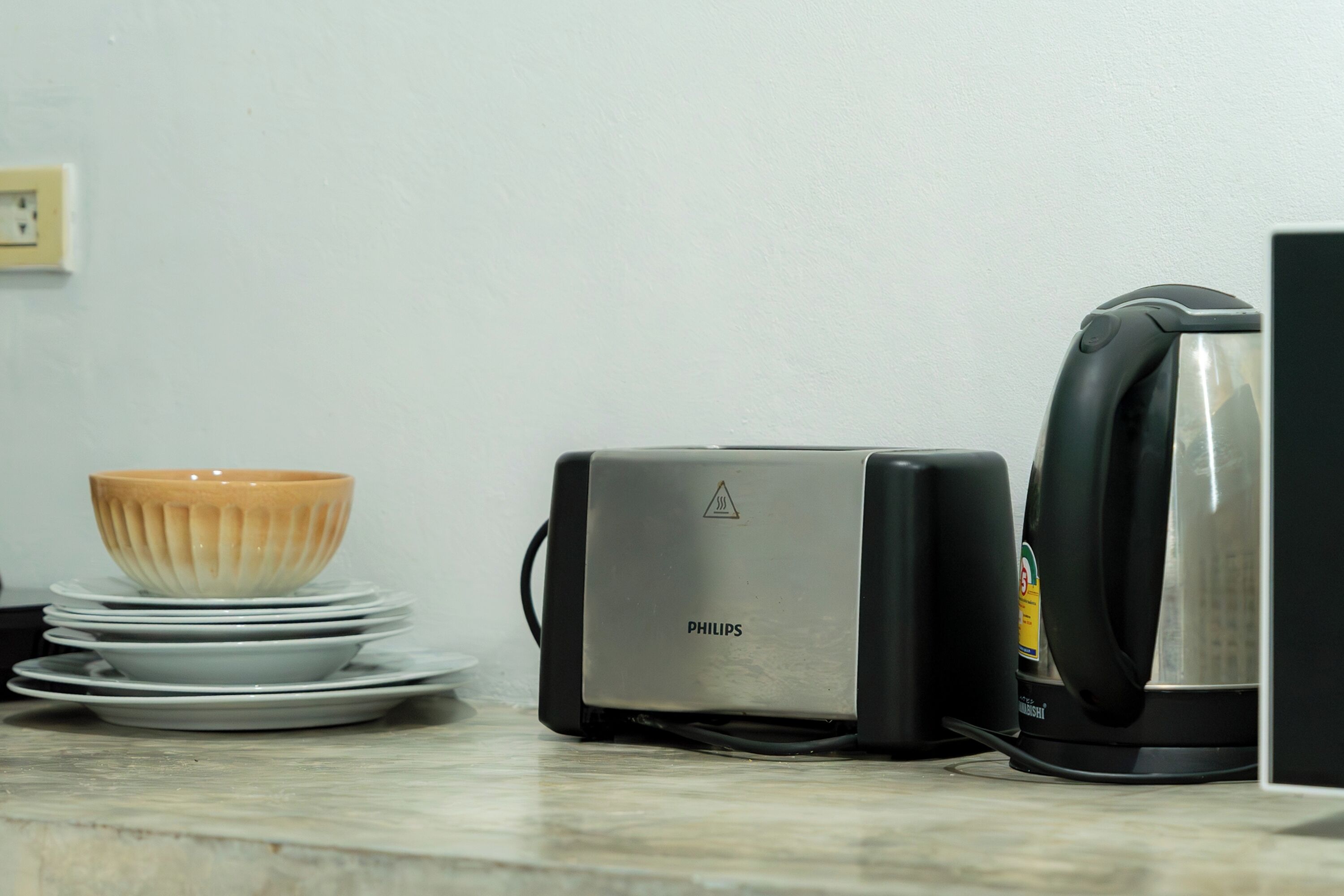 A kitchen counter with a stack of white plates, a bowl, a Philips toaster, and a kettle.