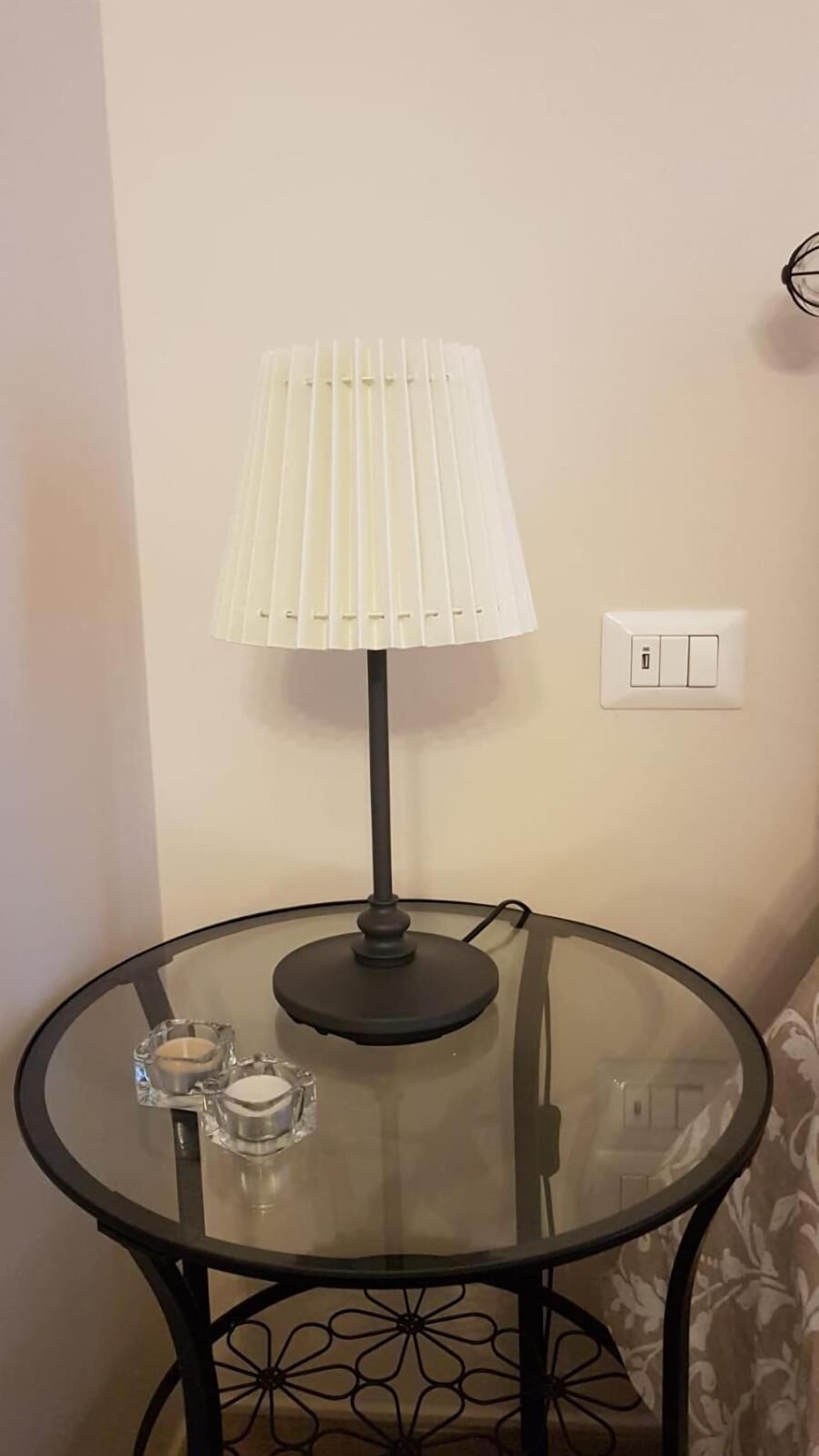 Uma mesa de vidro redonda com base de metal preta, uma luminária branca com cúpula franzida e uma tomada na parede.