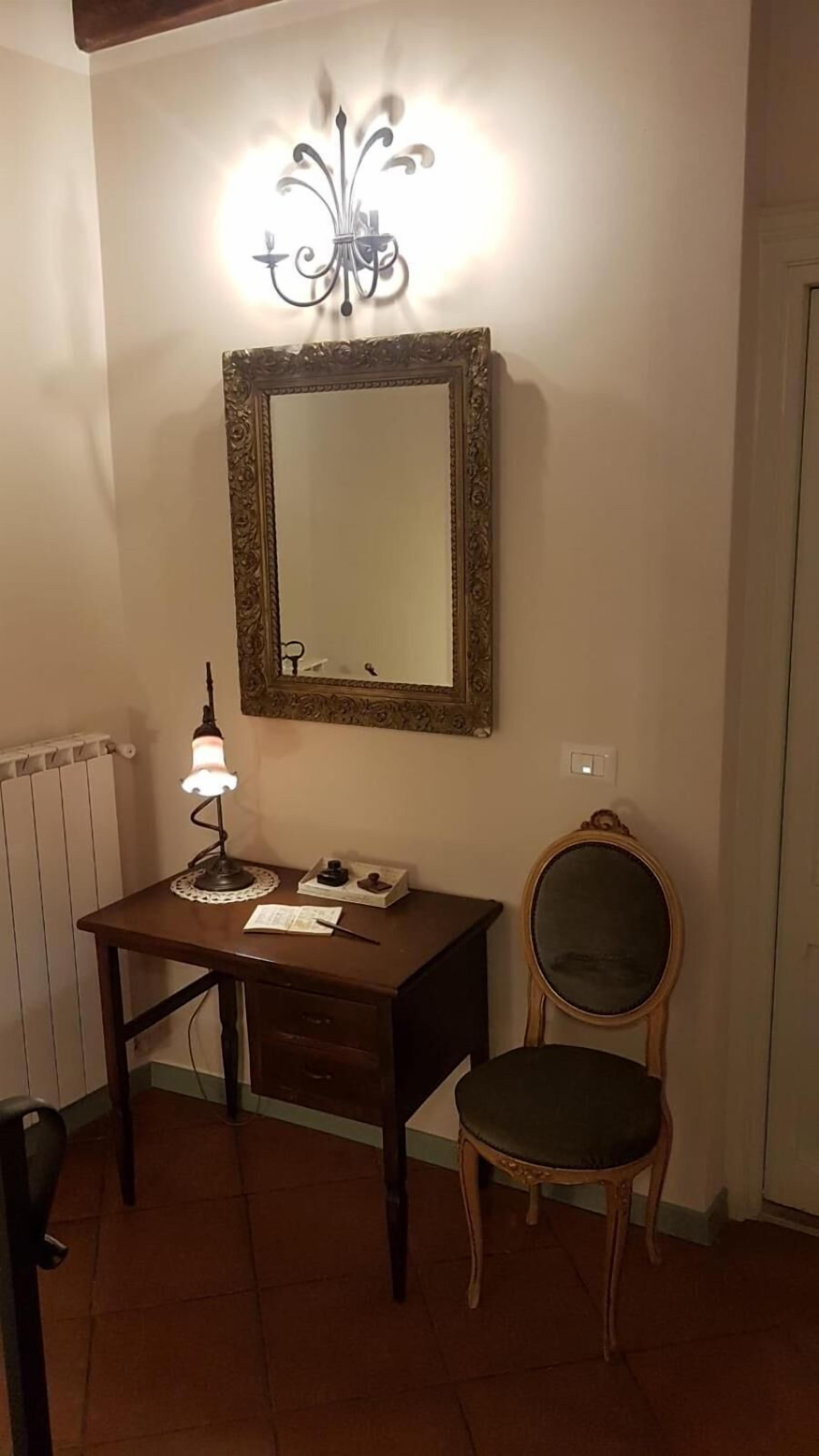 A room with a wooden desk, a chair, a wall-mounted mirror, and a wall sconce.