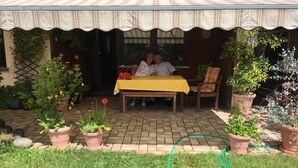 Une terrasse couverte avec une table et des chaises, entourée de plantes en pot et d’un tuyau d’arrosage.