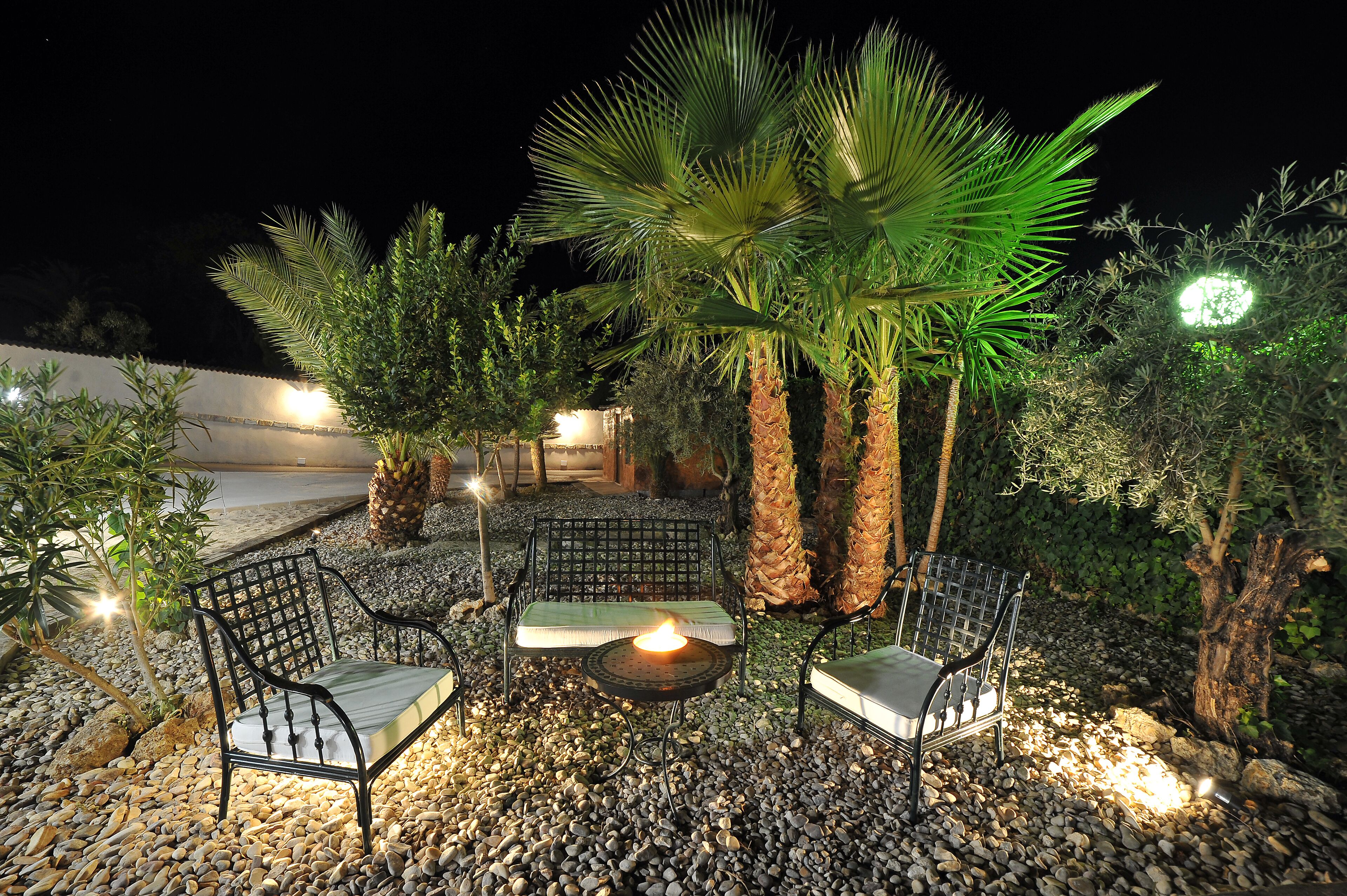 An outdoor seating area with two chairs and a table, surrounded by pebbles and palm trees, under night lighting.