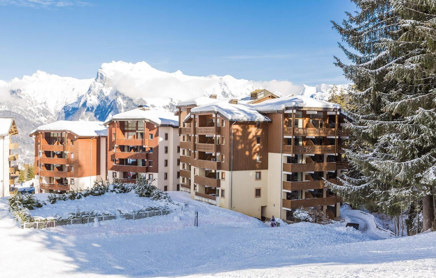 Un complejo turístico en la montaña cubierto de nieve, con edificios de varios pisos, balcones y una pista de esquí.