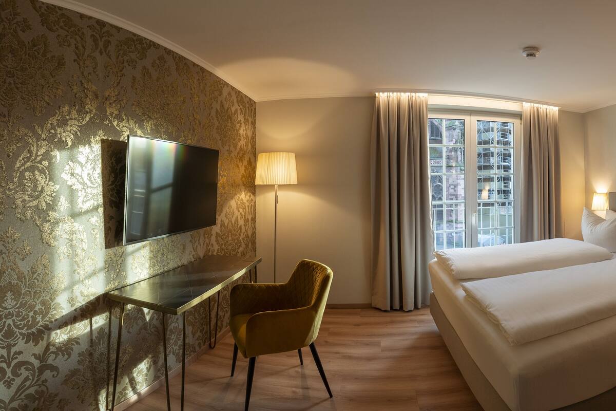 A hotel room with a bed, a desk, a chair, a TV, and a window with curtains.
