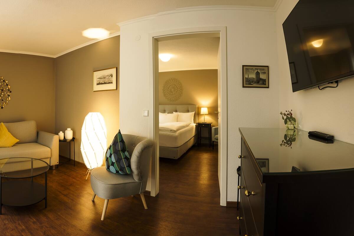 A modern living room with a sofa, armchair, and coffee table, a bedroom with a bed and bedside table, and a TV mounted on the wall.