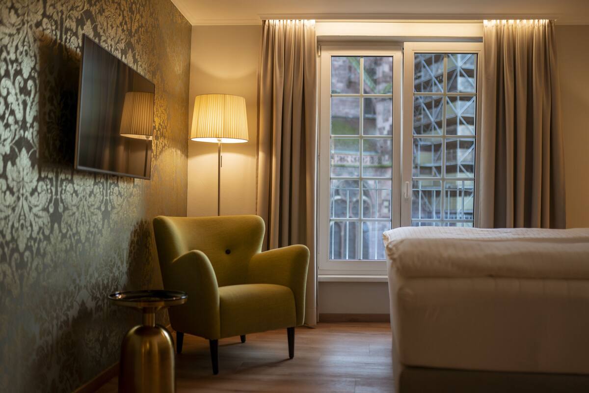 A hotel room with a bed, a green armchair, a wall-mounted TV, a floor lamp, and a window with curtains.