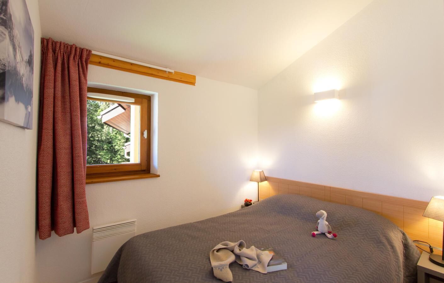 A hotel room with a bed, a window with curtains, a nightstand with a lamp, and a wall-mounted heater.