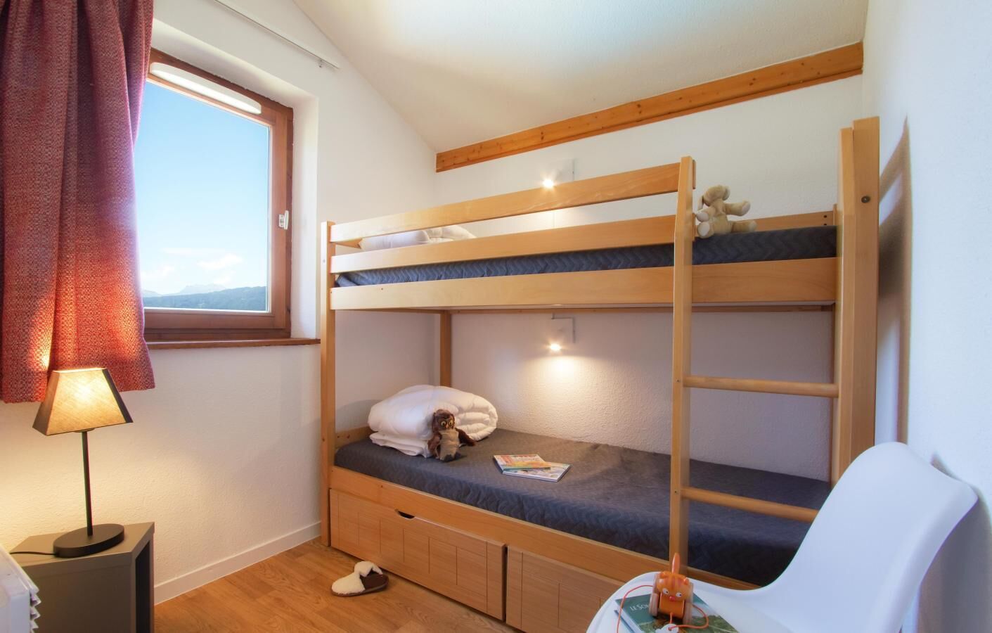 A bunk bed room with a window, a lamp, a chair, and a small table with a book.