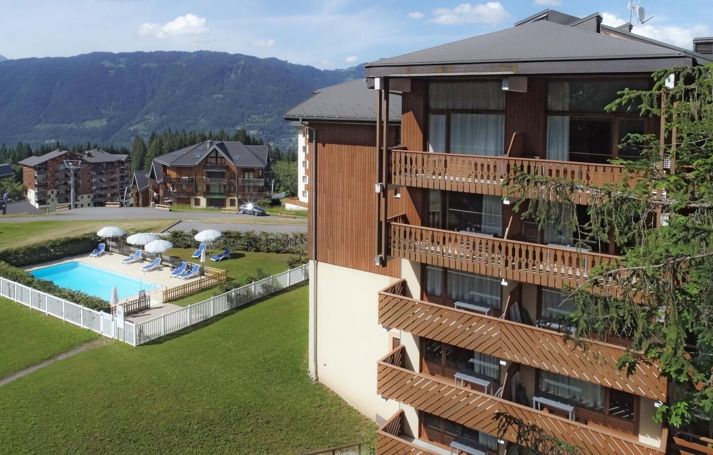 Un immeuble à plusieurs étages doté d’un espace piscine, comprenant des chaises longues et des parasols, le tout entouré d’une pelouse délimitée par une clôture.
