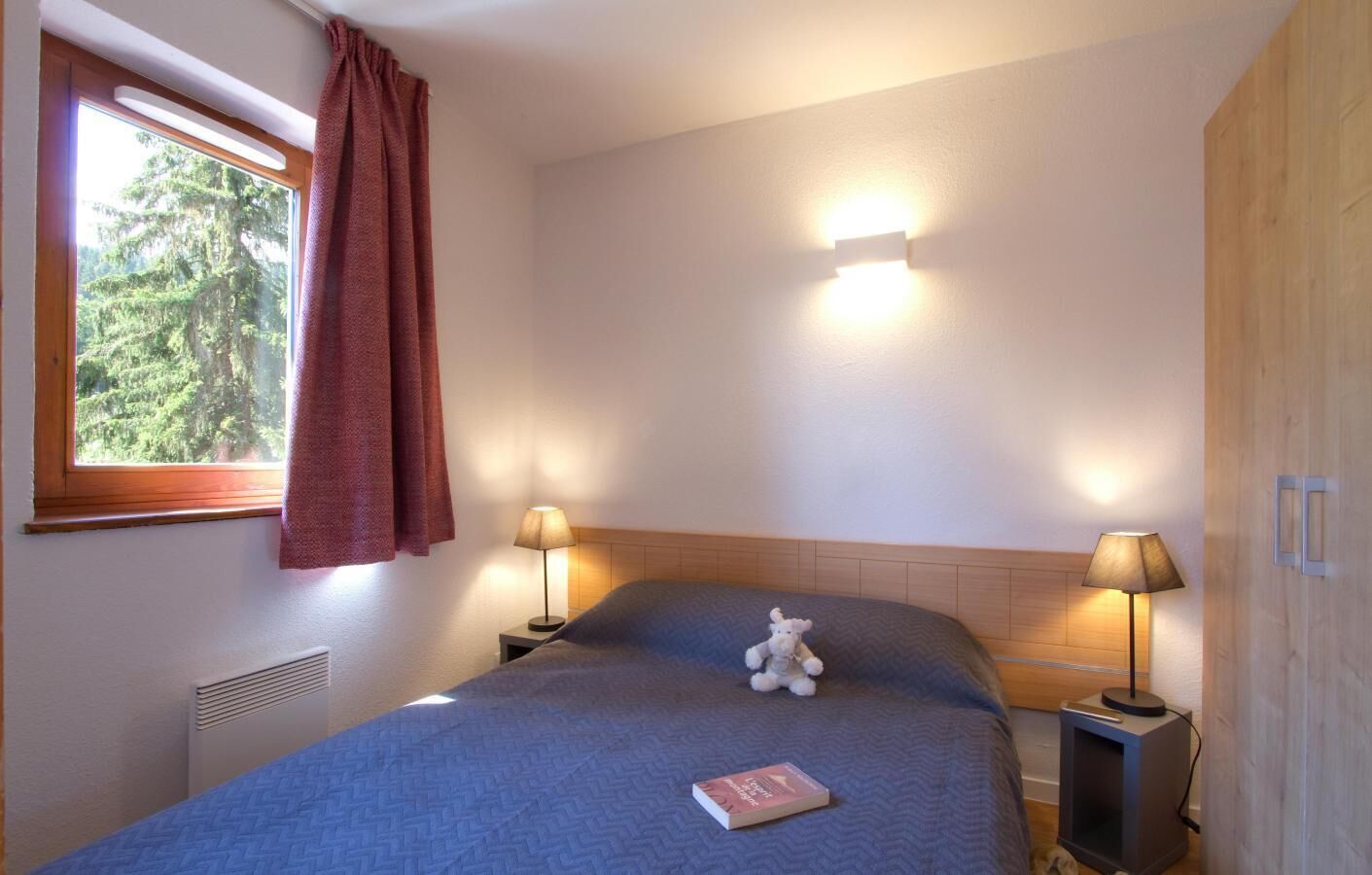 Ein Schlafzimmer mit Bett, zwei Nachttischlampen, einem Fenster mit Vorhängen, einer Wandleuchte und einem Heizkörper.