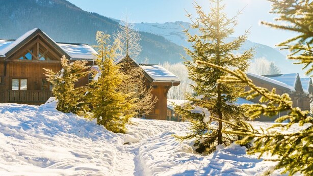 Une cabane en bois, dont le toit est recouvert de neige, entourée de neige et d’arbres, avec des montagnes en arrière-plan.