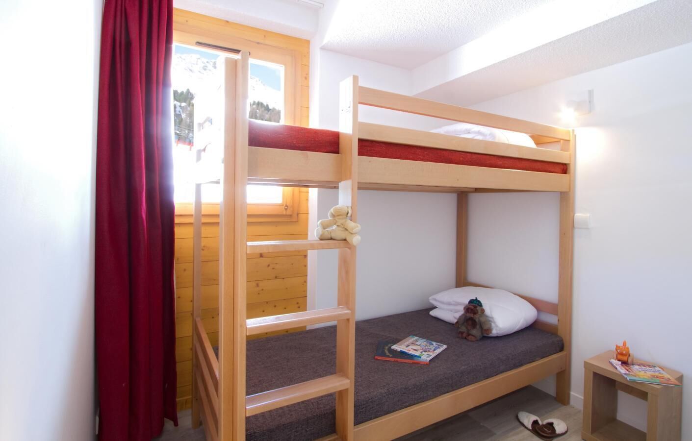 Une chambre avec un lit superposé, une fenêtre, une petite table et une paire de chaussons posés sur le sol.