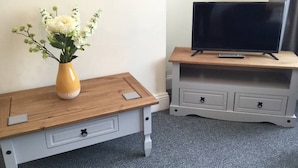A small wooden table with a vase of flowers, a television on a matching wooden stand, and a grey drawer unit. - Shaw Properties Teal Road (Darlington)