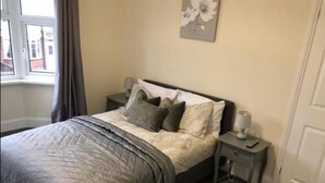 A neatly made bed with a grey quilt and pillows, a bedside table with a lamp, and a window with curtains. - Shaw Properties Teal Road (Darlington)