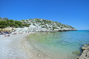 A pebble beach with clear water, a rocky hill, and umbrellas. - VILLA MADRAVE - FAMILIES AND FRIENDS (Pollensa)