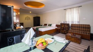 Una habitación de hotel con cama, sofá, mesa de comedor y una cesta de fruta, televisión y ventana con cortinas.
