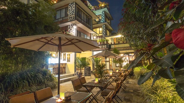 Un área de comedor al aire libre con mesas y sillas, rodeada de abundante vegetación y un edificio con varios balcones.