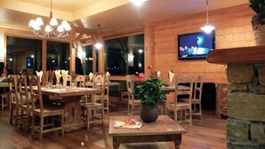 Un interior de madera con varias mesas y sillas para comer, un televisor montado en la pared y un gran ventanal con vistas al cielo nocturno.