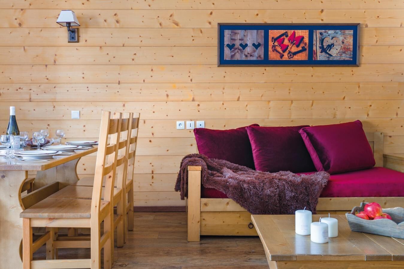 L’intérieur chaleureux d’un chalet avec une table à manger en bois, dressée pour deux personnes, un canapé avec des coussins rouges et une lampe fixée au mur.