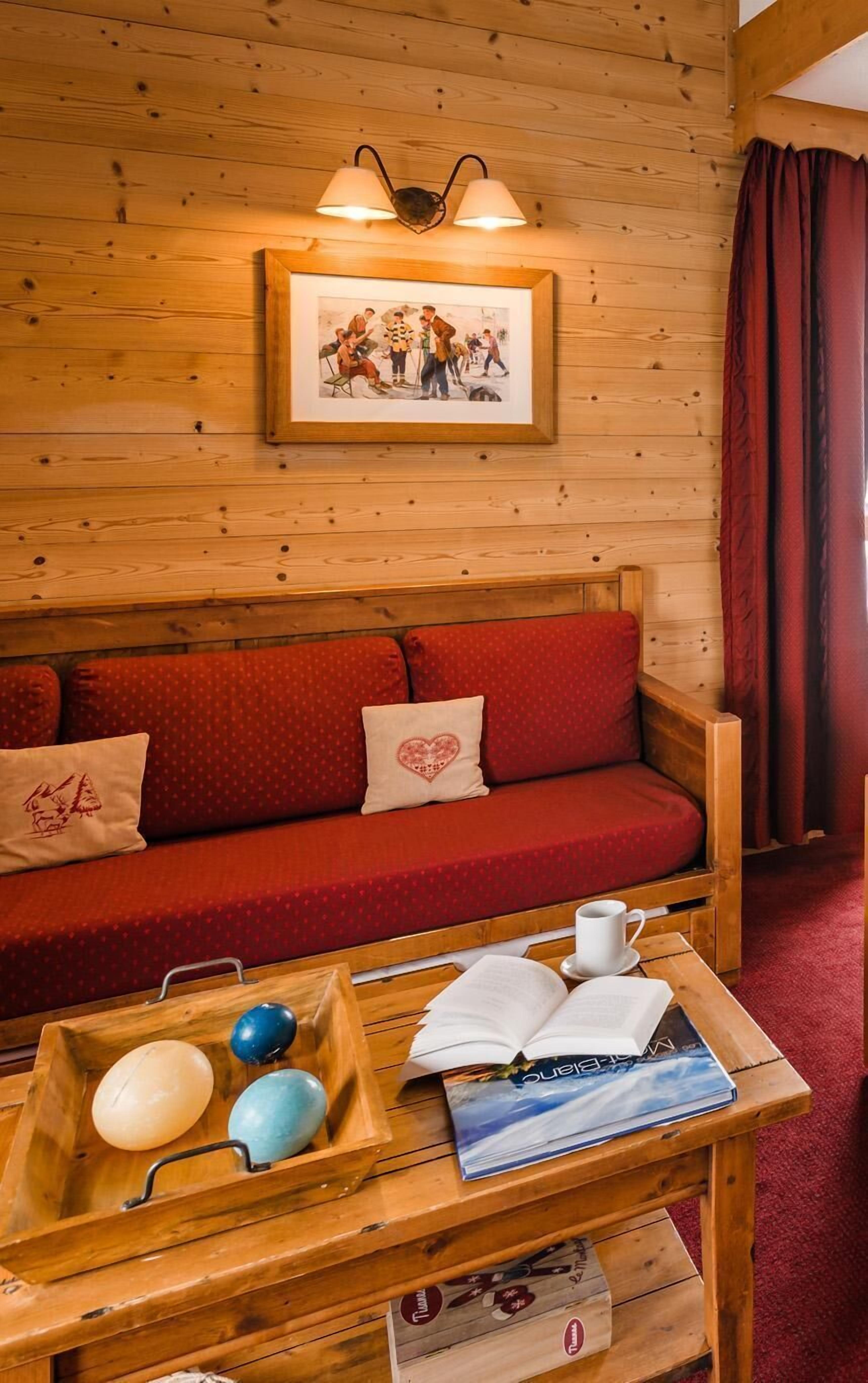 A cozy wooden cabin interior with a red sofa, a wooden table with a tray of eggs, and a framed picture on the wall.