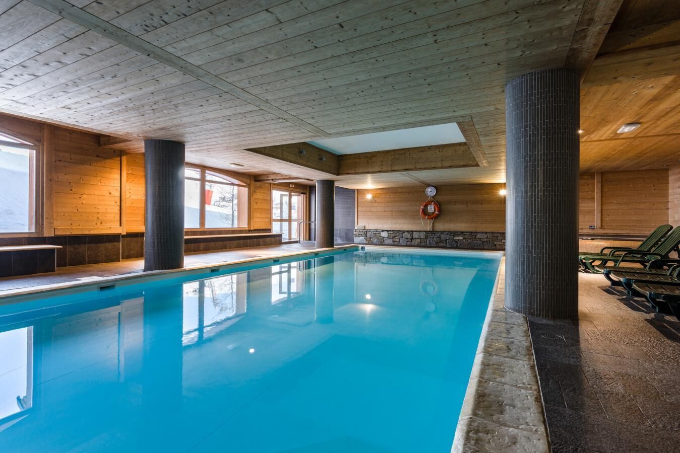 Une piscine intérieure aux murs et au plafond en bois, agrémentée d’éléments en pierre et de fauteuils de détente verts.