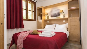 A bedroom with a wooden headboard, a red bedspread, white pillows, a wooden shelf with a painting, and a window with red curtains. - Wifi, 100m from shops, swimming pool, parking, tv, ski locker, 32m², Saint Sorlin d'Arves (Saint-Sorlin-d'Arves)