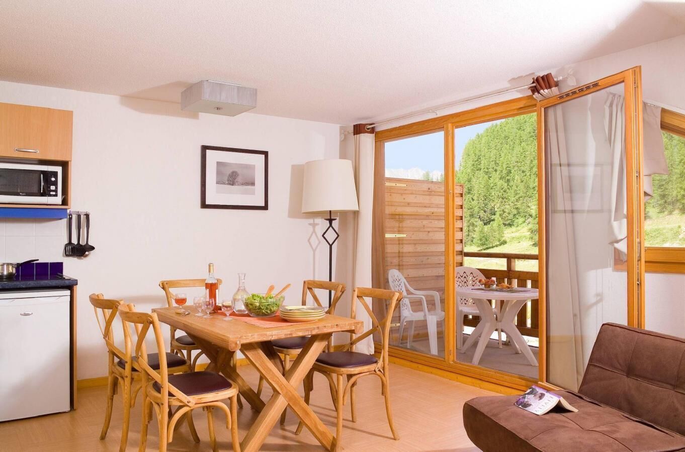 A cozy room with a wooden dining table, chairs, a sofa, a kitchen area with a microwave, and a large sliding glass door leading to an outdoor deck.