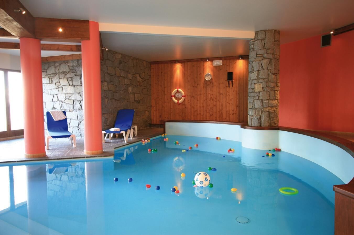 An indoor swimming pool with a stone wall and wooden ceiling, two blue lounge chairs, and colorful balls scattered in the water.