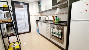 Eine Küche mit weißen Schränken, einer Mikrowelle, einem Herd und einem Kühlschrank. Ein Regal mit Küchenutensilien und ein Fenster mit Jalousien.