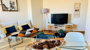 Ein modernes Wohnzimmer mit einem Glastisch, einer Couch mit gemusterten Kissen, einem Flachbildfernseher, einer kleinen Pflanze und einem dekorativen Schild an der Wand.