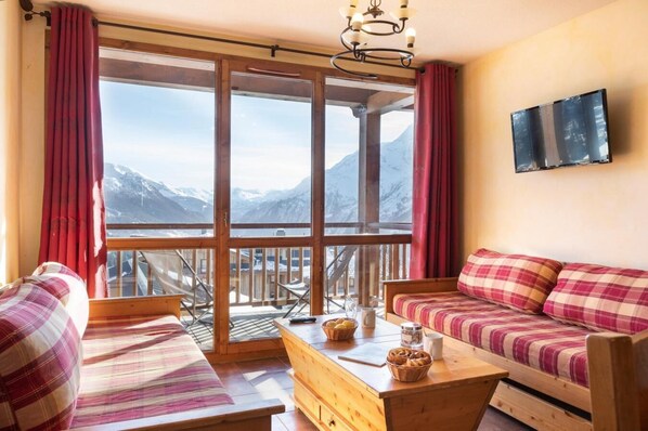 A cozy room with a wooden sofa and a bench, a wooden table with a basket and a mug, and a large window with a mountain view. - Wifi, ski-in/ski-out & foot of the slopes, swimming pool, sauna, hammam, balcony, tv, ski locker (La Rosière Montvalezan)