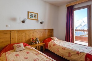 A hotel room with two beds, a wooden headboard, a small table with two teddy bears, a framed picture on the wall, and a window with a view of snow-capped mountains. - 200m from resort center, swimming pool, sauna, hammam, terrace, balcony, tv, ski locker, 47m² (Fontcouverte-la-Toussuire)