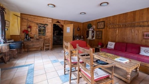 A cozy, rustic room with wooden walls and a tiled floor. It includes a dining table with chairs, a red sofa, and a small kitchen area with wooden cabinets and a sink.