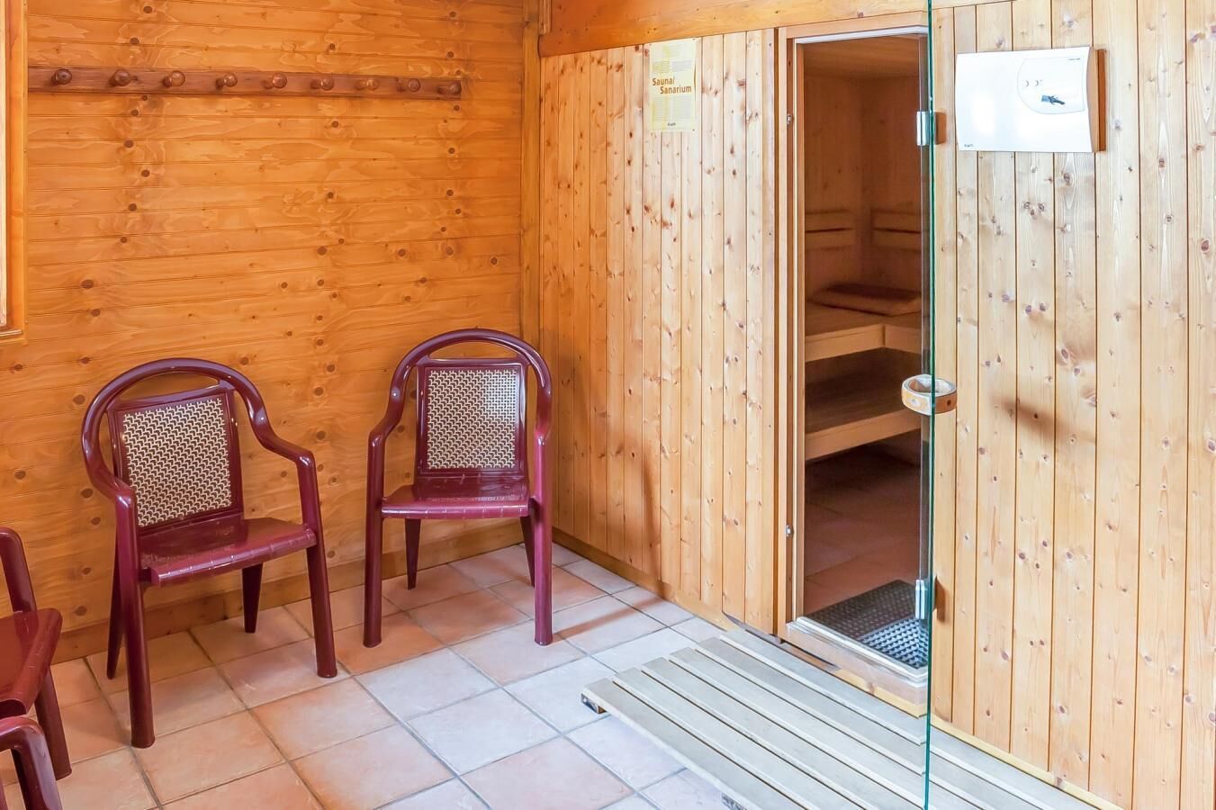 A sauna room with wooden walls and a glass door, two wooden chairs, and a tiled floor.