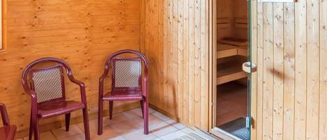 A sauna room with wooden walls and a glass door, two wooden chairs, and a tiled floor.