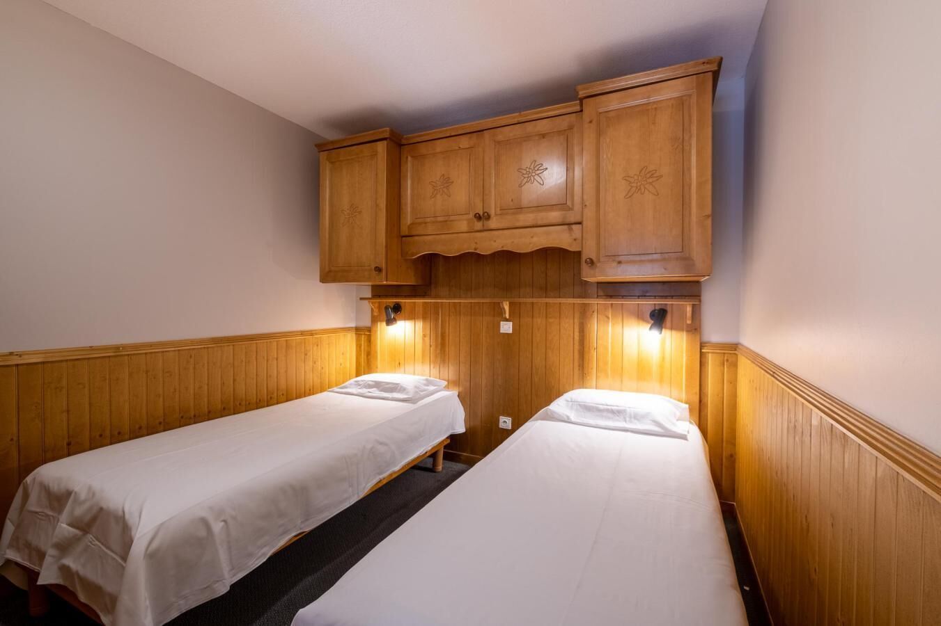 A small, single-room accommodation with two single beds, wooden paneling, and a wooden cabinet above the beds.