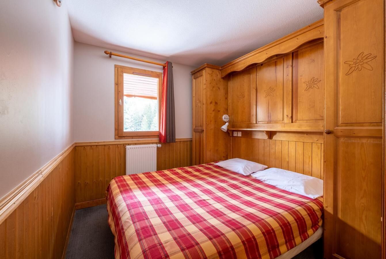 A bedroom with a plaid bedspread, wooden wardrobe, and a window with blinds.