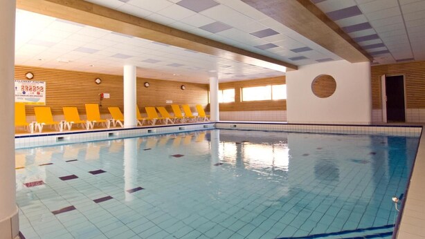 An indoor swimming pool with tiled floors and walls, yellow chairs along the side, and a wooden ceiling with recessed lighting.