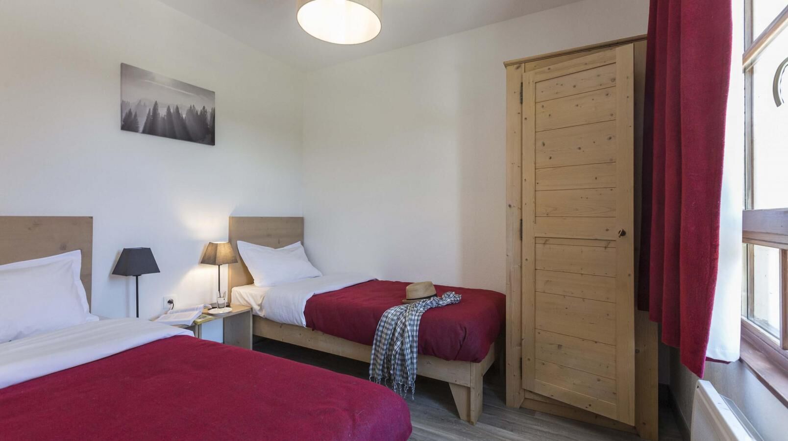 A hotel room with two beds, a wooden wardrobe, a nightstand with a lamp, and a painting on the wall.