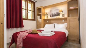 Un dormitorio con cabecera de madera, colcha roja, almohadas blancas, estante de madera con un cuadro y una ventana con cortinas rojas.